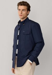 Jonge man draagt een marineblauwe jas met knopen over een gestreepte trui en beige broek, staat met één hand in de zak tegen een effen achtergrond.