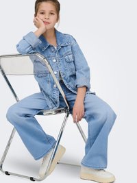 Chaqueta de mezclilla azul claro y jeans de pierna ancha, combinados con zapatillas blancas. La niña está sentada en una silla plegable cromada, apoyando la barbilla en su mano.