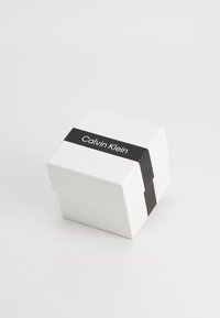 Caja de cartón blanca con una banda negra que presenta el texto "Calvin Klein". La caja tiene una forma rectangular simple y una textura suave.