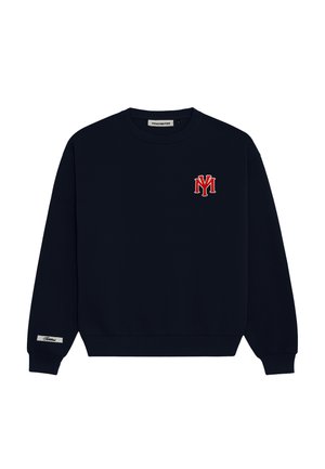 Ung person iført marineblå sweatshirt med NY-logo, hvide basketballshorts, hvide sokker, brune sko og en beige baseballkasket med design.
