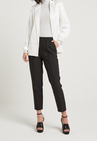 Weißes Satin-Blazer über einem glitzernden grauen Rollkragenpullover, kombiniert mit schwarzen tapered Hosen und schwarzen Plateauschuhen.