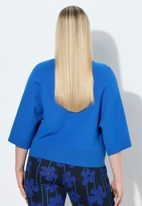 Blauer Kurzarmpullover mit geripptem Saum, kombiniert mit schwarzen Hosen mit großen blauen Blumenmustern. Weiche Stricktextur.