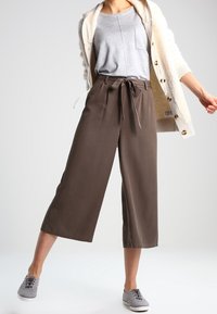 Bruine culottebroeken met een strik taille, gecombineerd met een grijze tanktop en een crèmekleurige cardigan. Model staat in casual grijze schoenen en toont een ontspannen pasvorm.