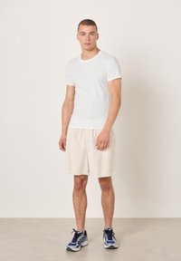 Maglietta bianca in cotone con collo rotondo, abbinata a pantaloncini beige chiaro testurizzati e scarpe da corsa con accenti blu e argento.