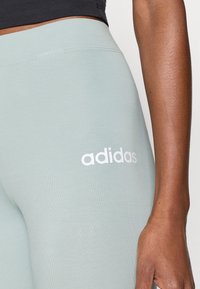 Svetlozeleni legingi izdelani iz raztegljive tkanine z gladko teksturo; vsebujejo bel logo "adidas" na stegnu.