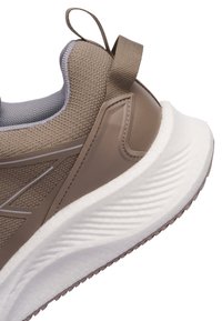 Detailní záběr na patu sportovní boty v béžové a taupe barvě s texturovanou tkaninou, poutkem a tlustou bílou polstrovanou podrážkou.