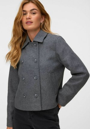 Femme aux longs cheveux blonds porte une veste grise en laine à double boutonnage et un pantalon noir, debout avec une main dans la poche, sur fond blanc.