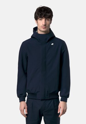 Uomo che indossa una giacca con cappuccio blu navy e pantaloni abbinati con un logo bianco sul petto sinistro, in piedi contro uno sfondo chiaro e uniforme.