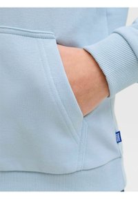 Sudadera azul claro con bolsillo canguro, puños acanalados y textura de tela suave. Detalles incluyen costuras cosidas y una etiqueta pequeña.