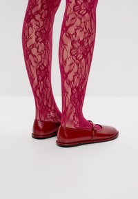 Medias de encaje rosa con un patrón floral y zapatos planos rojos brillantes con punta redondeada y detalle de correa, mostrados de perfil.
