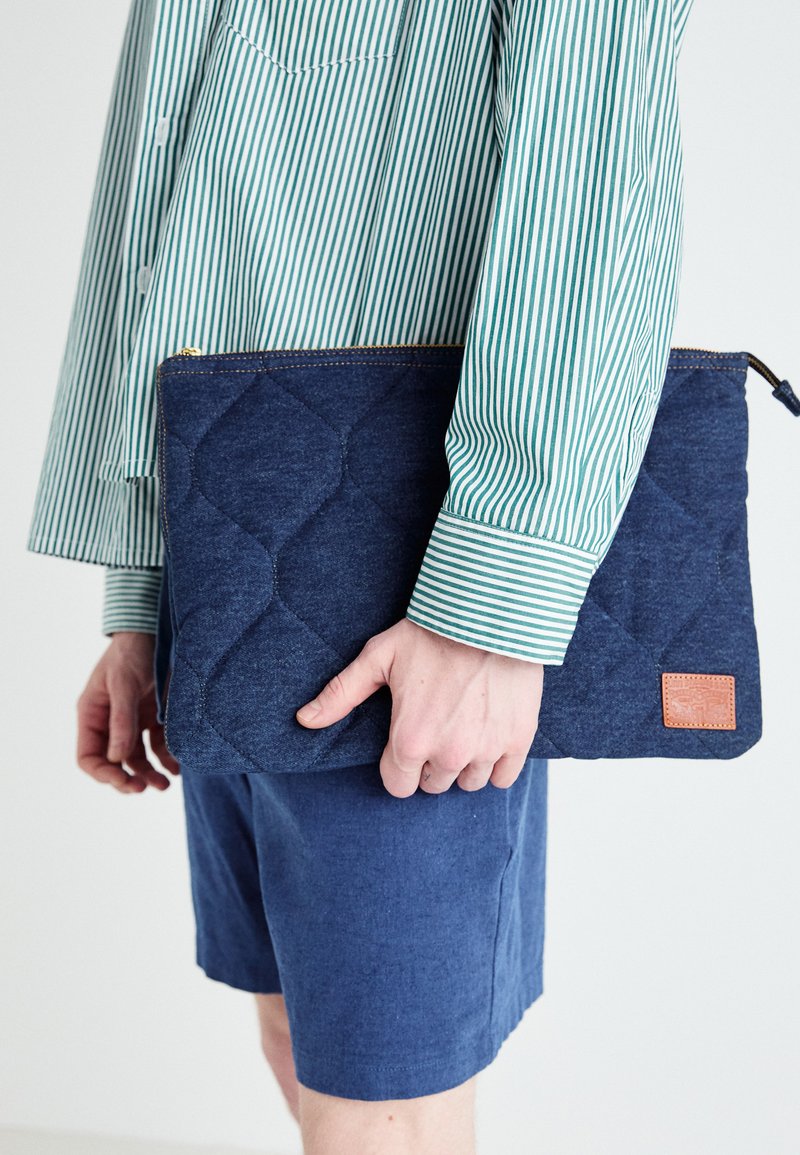 Denim laptop hoes, gewatteerd ontwerp, marineblauwe kleur, gouden rits, leren accentpatch. Vastgehouden door een persoon met een groen gestreept shirt.