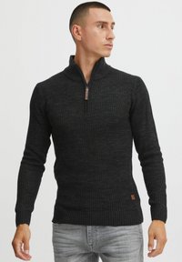 Svart zip-neck genser med ribbestruktur, lange ermer, og en brun lærlogo i hemmet, parret med lysegrå jeans.