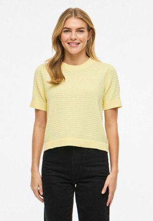 Femme souriante aux longs cheveux blonds portant un pull à manches courtes jaune clair texturé et un pantalon noir taille haute.