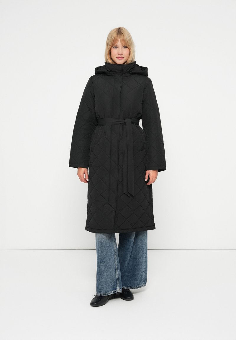 Juodas siuvinėtas ilgas paltas su kapišonu ir diržu, turintis plačias rankoves ir rombų rašt�ą. Derinamas su mėlynomis plačiomis kelnėmis ir juodomis batais.