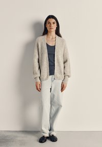 Svetlo sivi pleteni kardigan z rebrasto teksturo, odprtim sprednjim delom in spuščeno ramo, nošen preko temno barvnega majice brez rokavov in kombiniran s svetlo modrimi kavbojkami.