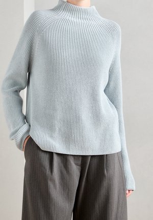 Personne portant un pull en maille côtelée bleu clair à manches longues et un pantalon large à rayures grises, les mains reposant le long du corps.