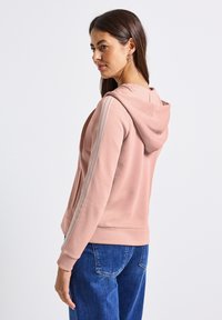 Felpa rosa con cappuccio regolabile, con strisce grigie lungo le maniche e una tasca a canguro. Indossata con jeans blu.
