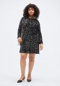 Fekete, flitteres ruha hosszú ujjakkal és kerek nyakkivágással, laza szabással, a térd felett végződik. Feket magas sarkú cipővel viselve.