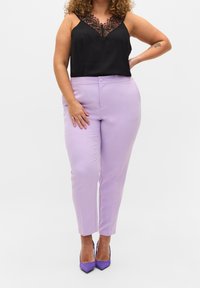 Kvinna klädd i svart spetskantad linne, högmidjade ljuslila byxor och lila klackar, står med en hand på höften mot vit bakgrund.