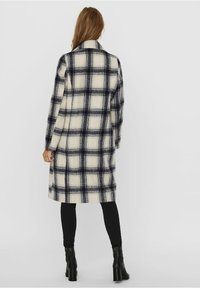 Rutet uldjakke i creme- og marineblå farver, lang model med krave, struktureret stof, sammen med sorte leggings og stiletter.