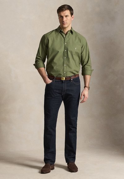 Muž stojaci s jednou rukou v vrecku, oblečený v olivovo zelenej košeli na gombíky, tmavých rifliach, hnedom opasku a hnedých topánkach na jednofarebnom pozadí.