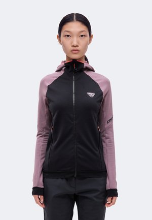 Un modèle se tient devant un fond uni, vêtu d’une veste à capuche noire et mauve, zippée, avec un pantalon noir assorti.