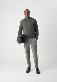 Pulover de tricot de culoare verde închis, cu guler cu jumătate de fermoar, asortat cu pantaloni cargo gri deschis. Se poartă pantofi negri și o geantă structurată neagră.