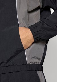 Hand in der Tasche einer schwarz-grau gestreiften Windbreaker-Jacke mit elastischen Bündchen und Saum, wobei ein Teil der passenden Hose sichtbar ist.