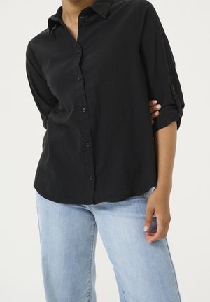 Frau trägt ein schwarzes Hemd mit Knöpfen, hochgekrempelten Ärmeln und hellblauen Jeans, steht mit einer Hand in der Taille.