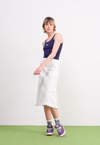 Marine tank top, hvit ankellang skjørt med volanger, lilla joggesko og svart-hvite rutete sokker på en lys grønn gulv.