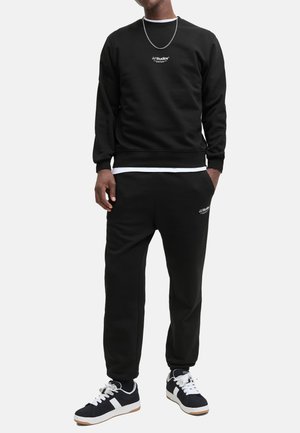 Black sweatshirt and sweatpants set with ribbed cuffs, featuring a small white logo on the chest and leg. Paired with black and white sneakers.