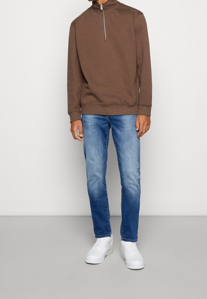 Sweat-shirt marron à col zippé associé à un jean bleu clair et des baskets blanches ; coupe décontractée avec poignets et ourlet côtelés.