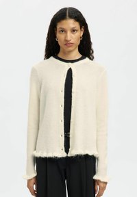 Woman with curly dark hair wears a cream fuzzy button-up cardigan over a black top with black pants, standing against a plain white background.