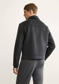 Chaqueta de mezcla de lana gris con un cuello estructurado, puños con botón y una silueta corta. Presenta costuras visibles y aberturas laterales.