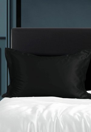 Ein schwarzes rechteckiges Kissen lehnt an einem dunkel gepolsterten Kopfteil mit weißem Satinsbetttuch darunter in einem minimalistischen Schlafzimmer.
