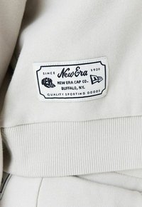 Tessuto grigio chiaro con orlo rigato, presenta un'etichetta bianca con testo nero: "New Era Cap Co., Buffalo, NY, Quality Sporting Goods."