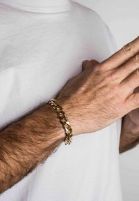 Kuldne kettbracelet, mis on valmistatud metallist ja millel on omavahel seotud lingid. Pind on sile ja läikiv, kantud randmel, millel on kerge karv.