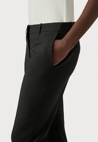 Main à la peau foncée reposant dans la poche avant d'un pantalon noir sur mesure porté avec un haut blanc rentré, montrée de la taille à la mi-cuisse.