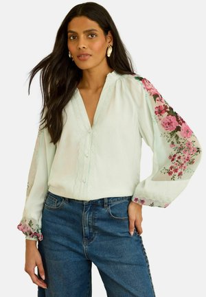 Frau mit langen dunklen Haaren, die eine helle Bluse mit transparenten floralen Ärmeln und blaue Jeans trägt, dazu goldene Ohrringe.