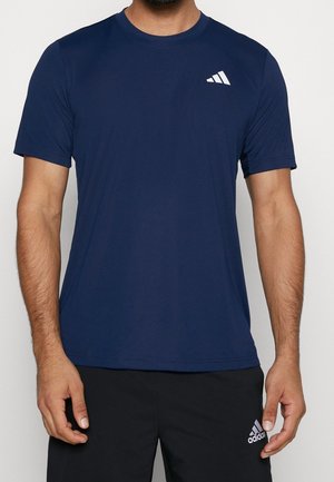 Mężczyzna ubrany w granatowy t-shirt Adidas i czarne spodenki Adidas, stojący na jednolitym jasnym tle.