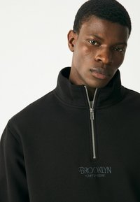 Black zip-up sweatshirt with high collar, featuring "BROOKLYN FORT GREENE" embroidered in blue. Smooth texture and modern cut.
