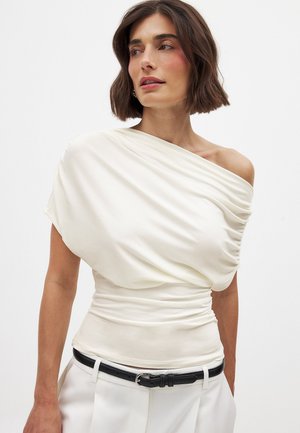 Off-white gedrapeerde top met asymmetrische halslijn, geplooide textuur en korte mouwen, gecombineerd met een zwarte riem en lichtgekleurde broeken.