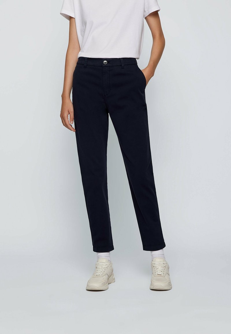 Mörkblå byxor i skräddarsydd passform, tillverkade av ett slätt tyg. Har en framknäppning med knapp och sidofickor, i kombination med vita sneakers.