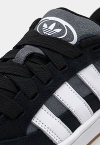 Svarta mockaskor med grå accenter, med vita läderstripes och en rund logotyp på tungan. Texturerade snören och sömdekor.