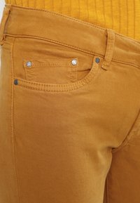 Mustard yellow denim pants with a smooth texture, five-pocket design, and silver hardware accents on pockets and waistband.