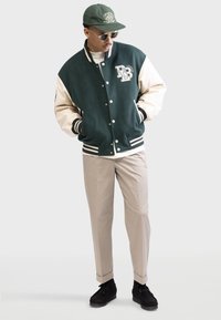 Green and cream varsity jacket with ribbed cuffs, featuring logo on the chest. Light beige trousers and black slip-on shoes complete the look.