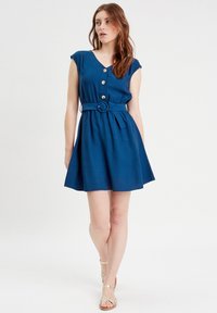 Dunkelblaues Kleid mit kurzen Ärmeln, V-Ausschnitt, Knopfdetail und einem taillierten Bund. A-Linien-Silhouette, der Stoff wirkt leicht und glatt.