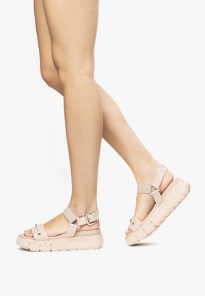 Béžové platformové sandály s nastavitelnémi kotníkovými proužky, zdobené cvočky a texturovanými podrážkami. Design má robustní a strukturovaný tvar.