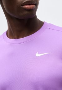 Κοντινό πλάνο ενός ατόμου που φοράει πουκάμισο λιλά με λευκό λογότυπο Nike στη στολή, εμφανίζοντας το κάτω μέρος του προσώπου και τον λαιμό.