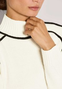 Frau trägt einen weißen Strickpullover mit schwarzem Saum, Hand auf dem Kinn, Lippen leicht geöffnet, neutraler heller Hintergrund.